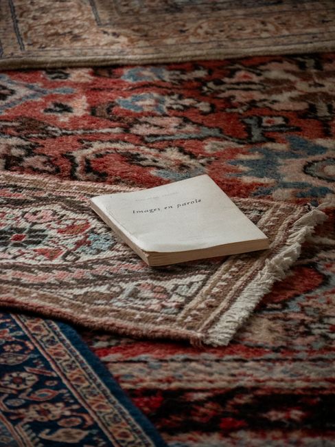A man in a gray long-sleeve shirt and dark trousers uses a vacuum cleaner to deep clean a living room with wooden herringbone floors. The room features large windows with sheer white curtains, allowin