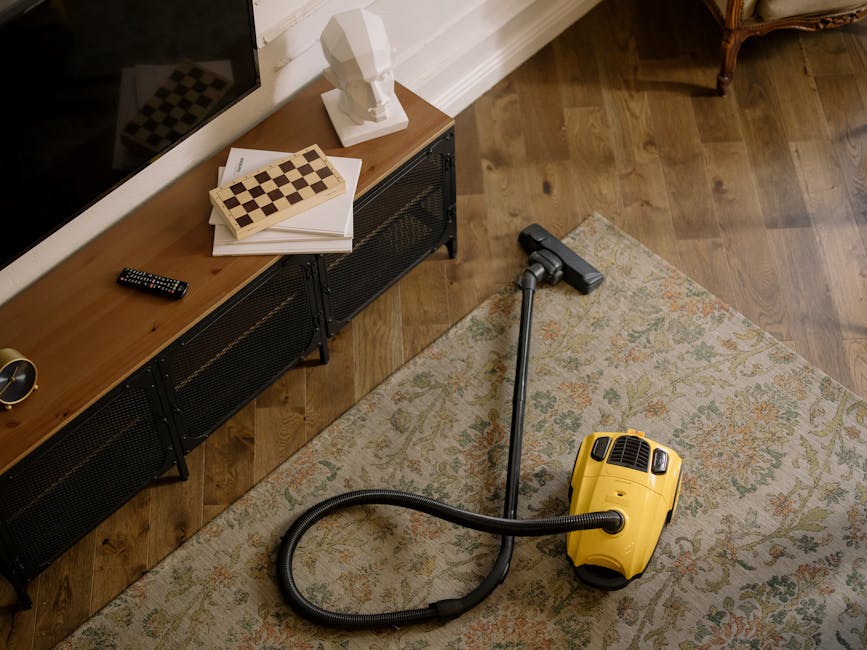 A person using a vacuum cleaner with a hose attachment to deep clean a patterned area rug in a domestic living space. The person is wearing casual beige shoes and grey jeans, standing on the rug which